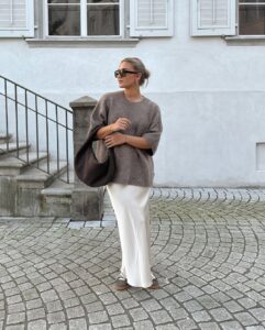 Mulher jovem de pele clara. Ela usa suéter marrom, saia acetinada off-white e tênis marrom claro
