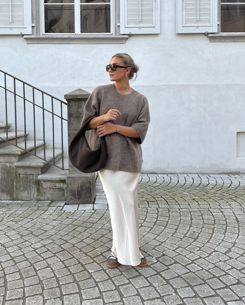Mulher jovem de pele clara. Ela usa suéter marrom, saia acetinada off-white e tênis marrom claro