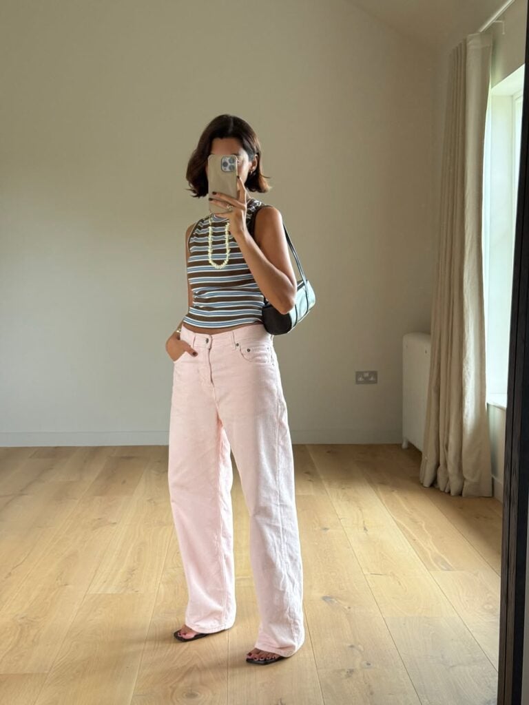 Mulher jovem, magra e com pele clara. Ela usa regata listrada, calça branca e chinelo de dedo