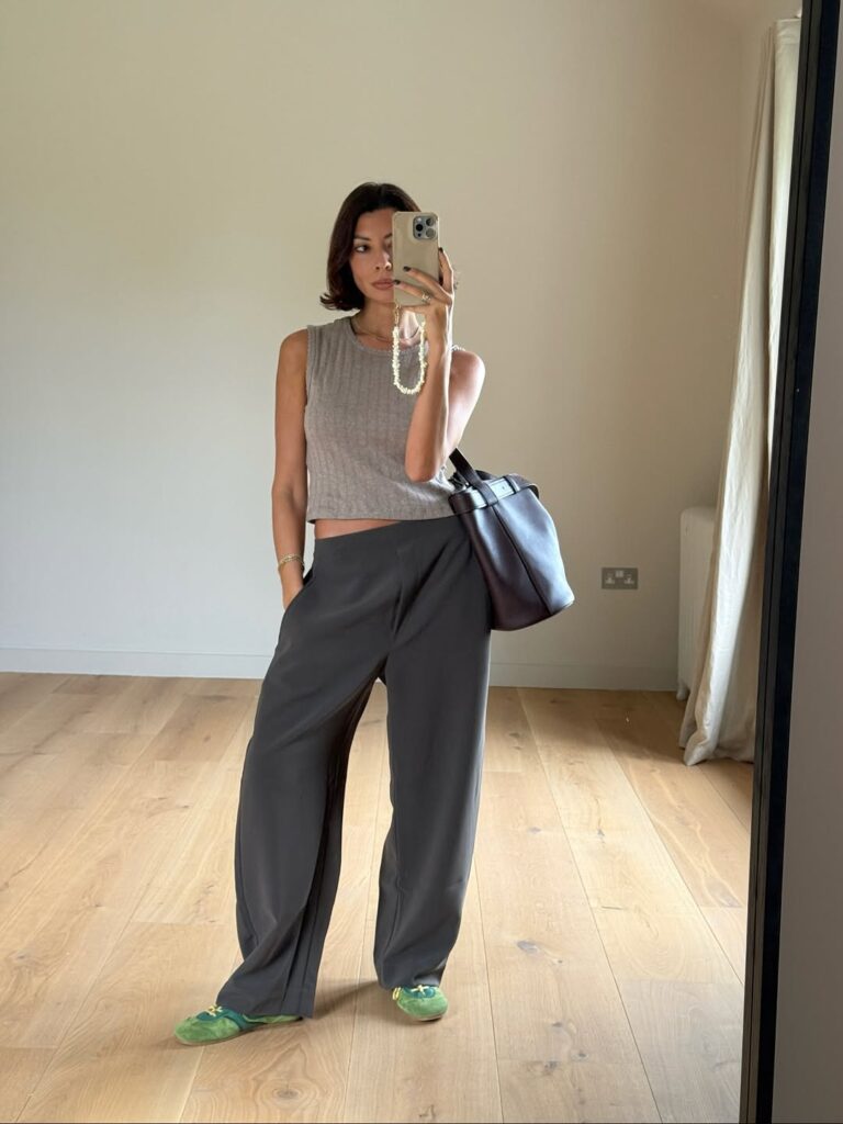 Mulher jovem, magra e com pele clara. Ela usa regata cinza claro, calça de alfaiataria cinza escuro e tênis verde