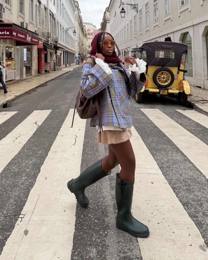 Mulher jovem e negra com cabelos longos. Ela usa jaqueta, saia e galochas