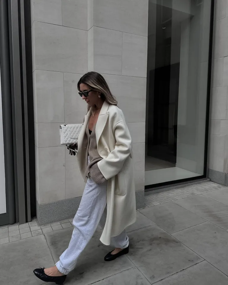 Mulher jovem e magra de pele clara. Eça usa casaco off-white longo, suéter marrom por baixo, calça branca e sapatilha preta