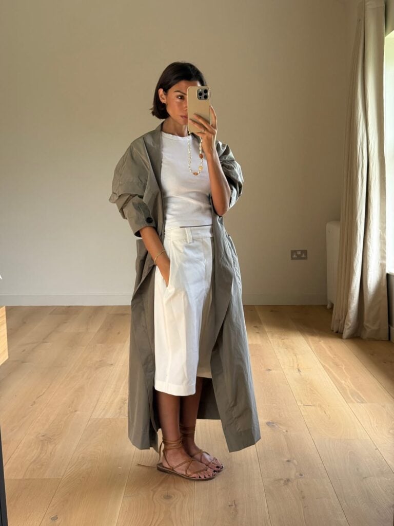 Mulher jovem e magra com pele clara. Ela usa trench coat, bermuda, camiseta e sandália rasteira com amarração.