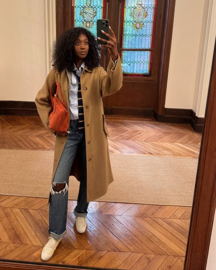 Mulher jovem, magra e de pele negra. Ela usa trench coat, camisa, gravata, calça jeans e tênis