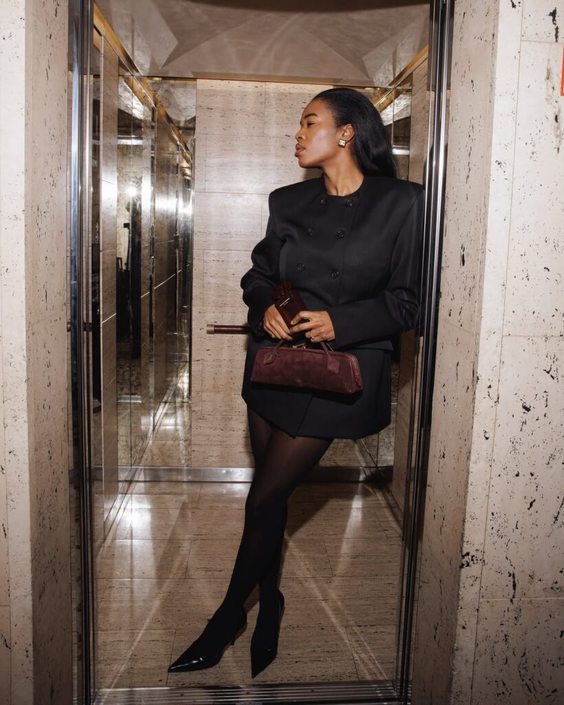 Mulher jovem, magra e de pele preta. Ela usa look all black com blazer, meia calça e salto alto de bico fino.