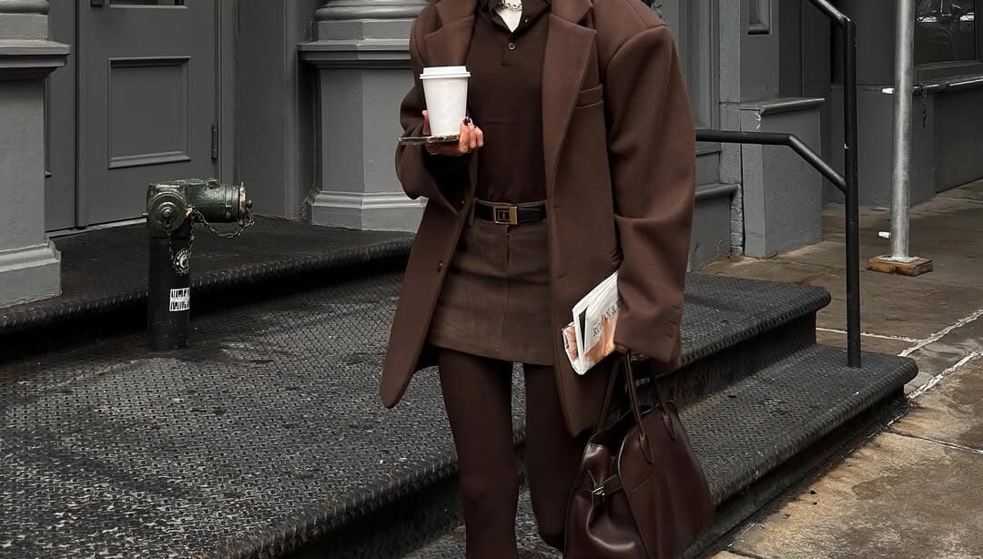 Mulher jovem, magra e de pele clara com cabelos curtos. Ela usa look marrom com blazer, csmids polo, minissaia de alfaiataria, meia-calça e mocassim
