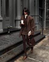 Mulher jovem, magra e de pele clara com cabelos curtos. Ela usa look marrom com blazer, csmids polo, minissaia de alfaiataria, meia-calça e mocassim