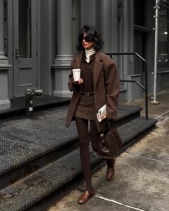 Mulher jovem, magra e de pele clara com cabelos curtos. Ela usa look marrom com blazer, csmids polo, minissaia de alfaiataria, meia-calça e mocassim
