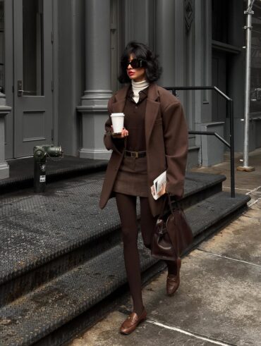 Mulher jovem, magra e de pele clara com cabelos curtos. Ela usa look marrom com blazer, csmids polo, minissaia de alfaiataria, meia-calça e mocassim