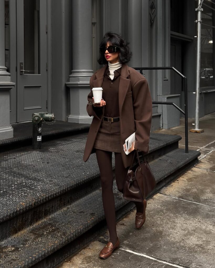 Mulher jovem, magra e de pele clara com cabelos curtos. Ela usa look marrom com blazer, csmids polo, minissaia de alfaiataria, meia-calça e mocassim