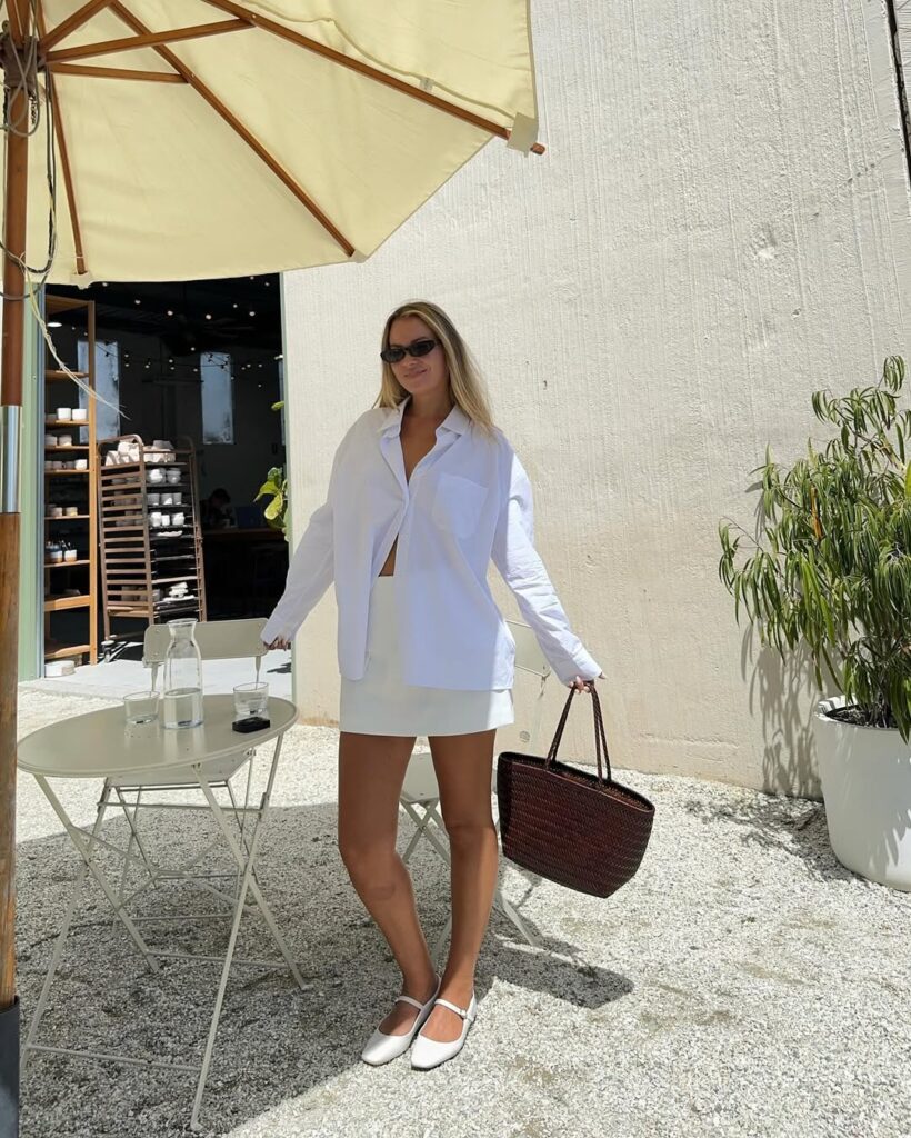 Mulher jovem, magra e de pele clara. Ela usa óculos de sol, camisa branca, saia branca, sapatilha branca e bolsa marrom média.