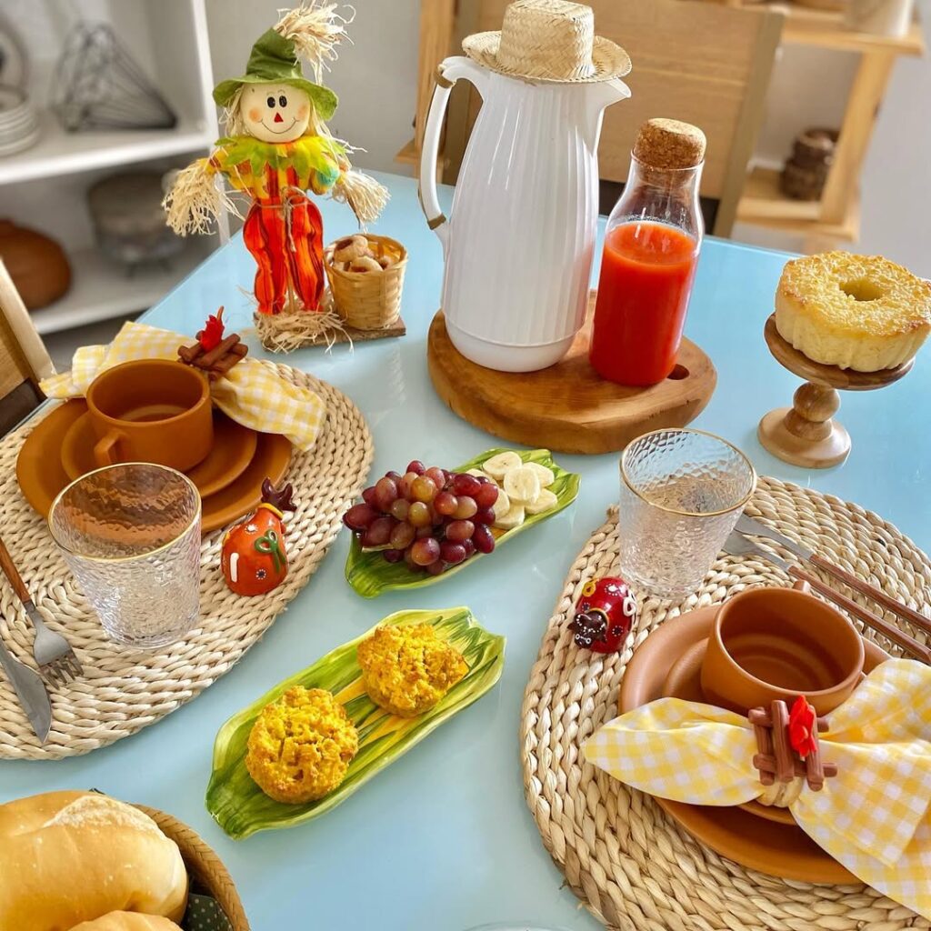 Mesa azul com café da manhã junino, sousplats de palha, louças marrom, guardanapos xadrez e espantalho decorativo