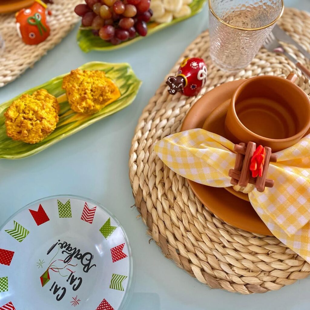 Mesa posta com sousplat de palha, prato e xícara marrom, guardanapo xadrez com mini fogueira