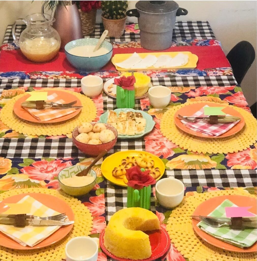Mesa posta junina com toalha xadrez, jogos americanos com tecido de chita, sousplats de crochê amarelo, pratos laranja e guardanapos xadrezes
