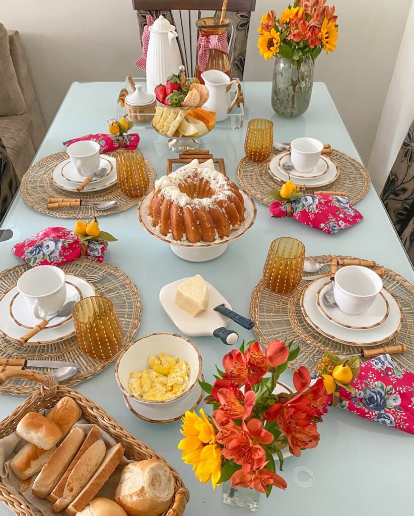 Ideia de café da manhã junino com queijo, pães, ovos mexidos e bolo, e guardanapos rosa com flores azuis