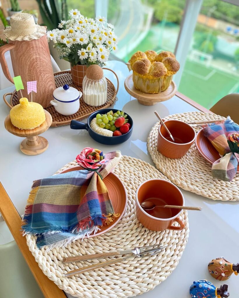 Mesa posta com sousplats de palha, louças marrom, guardanapos xadrezes, cuscuz decorado com bandeirinhas e garrafa térmica com chapéu de palha