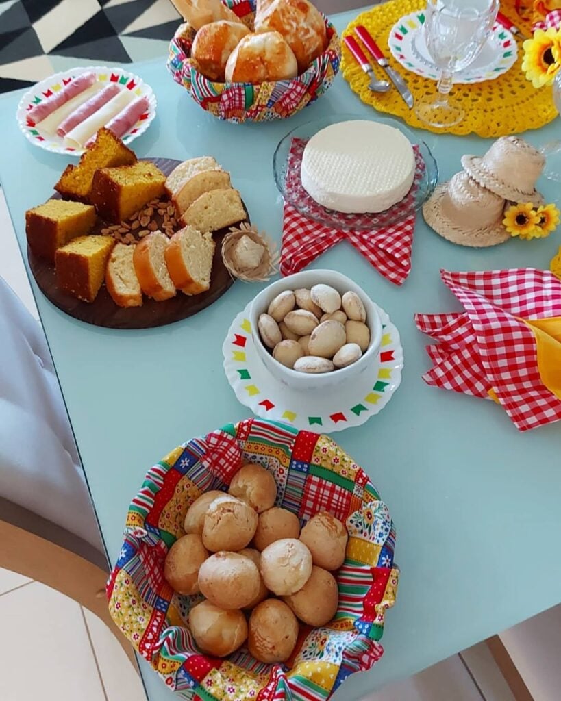 Mesa posta junia com tecidos xadrezes, chapéus de palha decorativos, pães de queijo, broas e bolos