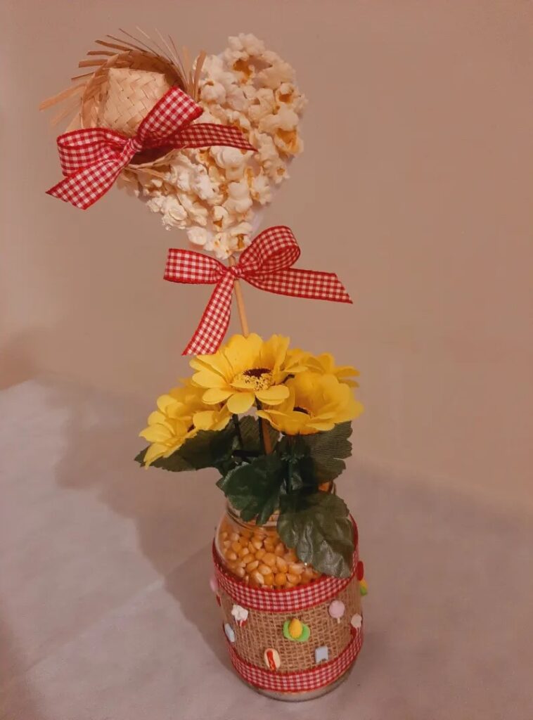 Centro de mesa junino com vidro decorado com juta e tecido xadrez, flores amarelas e coração feito com pipoca