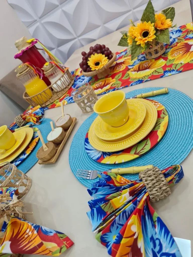 Mesa posta de São João com sousplats azuis, louças amarelas e caminho de mesa com flores