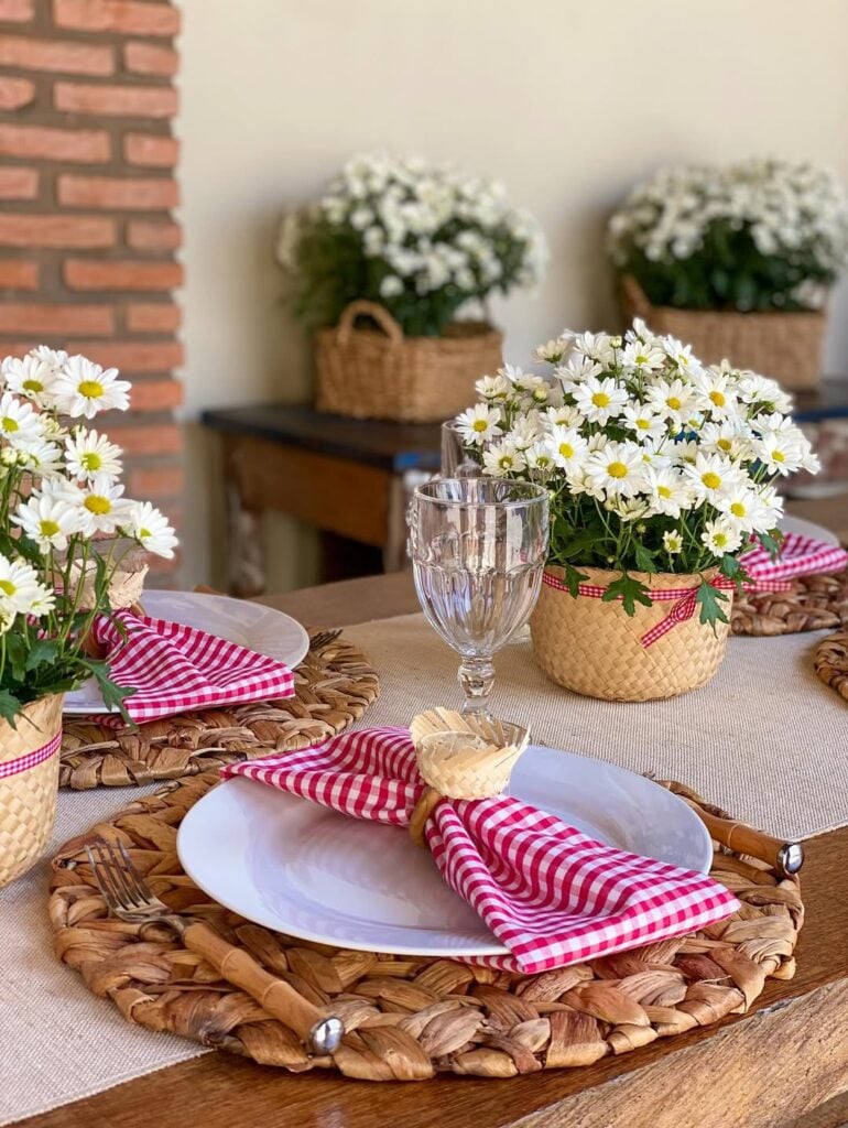 Mesa posta com sousplats de palha, pratos brancos, guardanapos xadrez em rosa e branco com chapéuzinho de palha e arranjos com margaridas