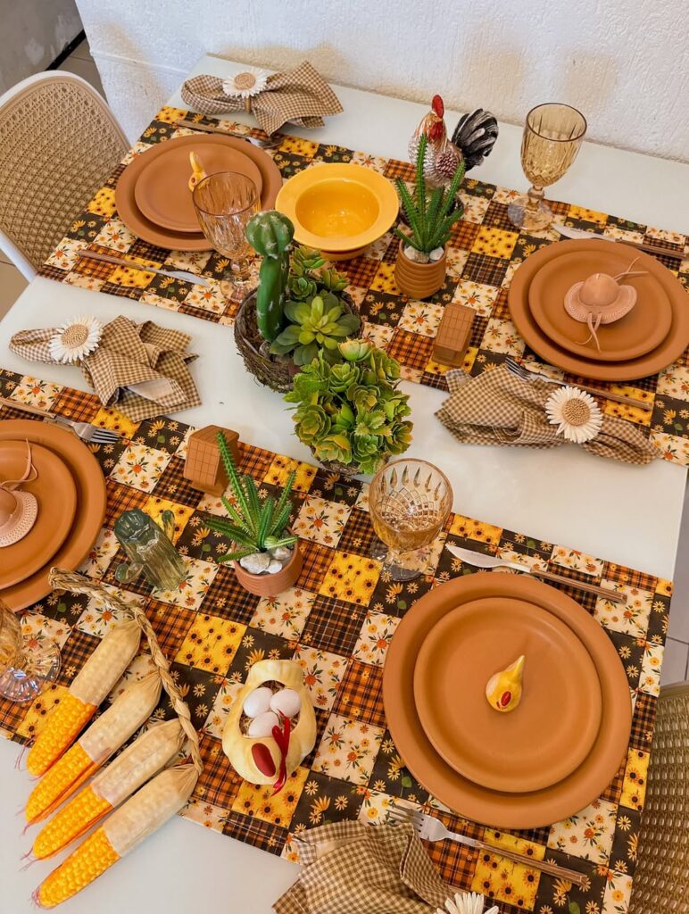 Mesa posta de São João com caminhos de mesa xadrez em tons de marrom e amarelo, louças marrom e enfeites como cactus e espigas de milho