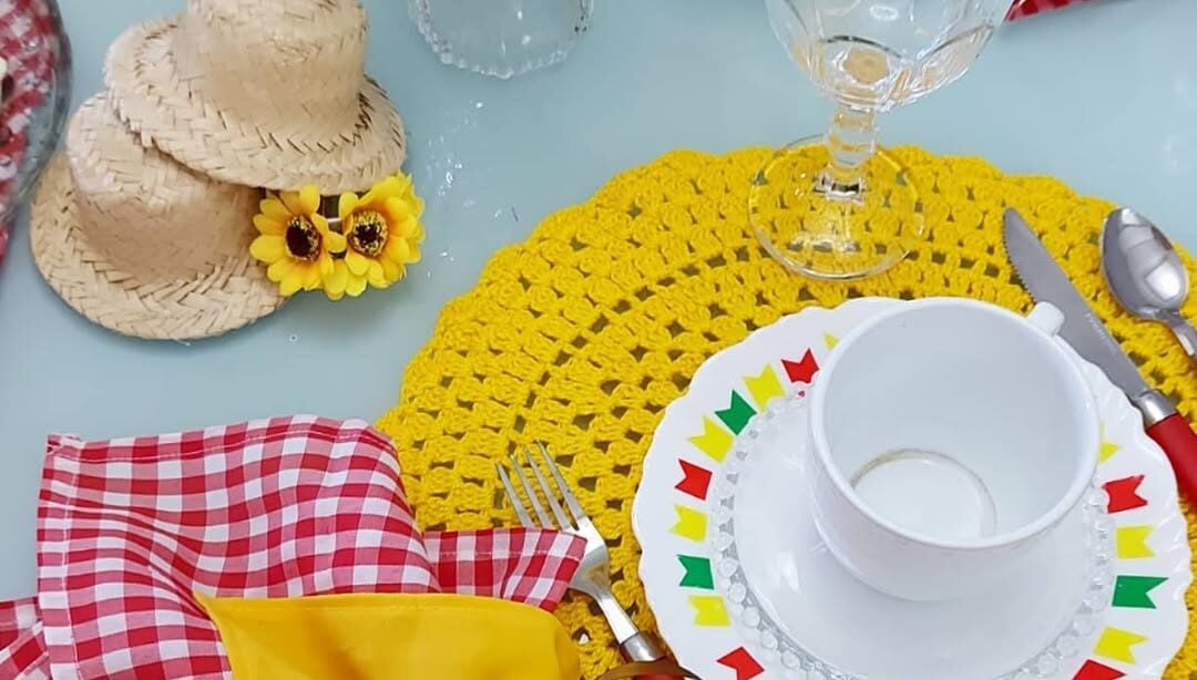 Mesa posta de São João com sousplats de crochê amarelo, prato branco com bandeirinhas coloridas e guardanapo xadrez com chapéuzinho de palha
