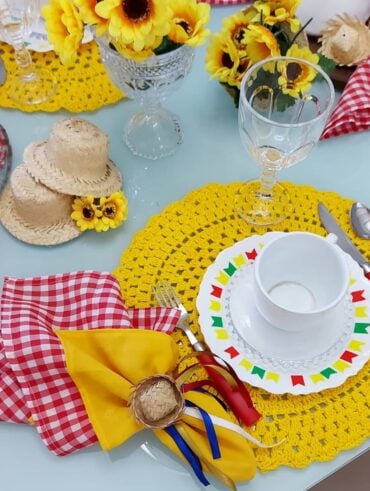 Mesa posta de São João com sousplats de crochê amarelo, prato branco com bandeirinhas coloridas e guardanapo xadrez com chapéuzinho de palha
