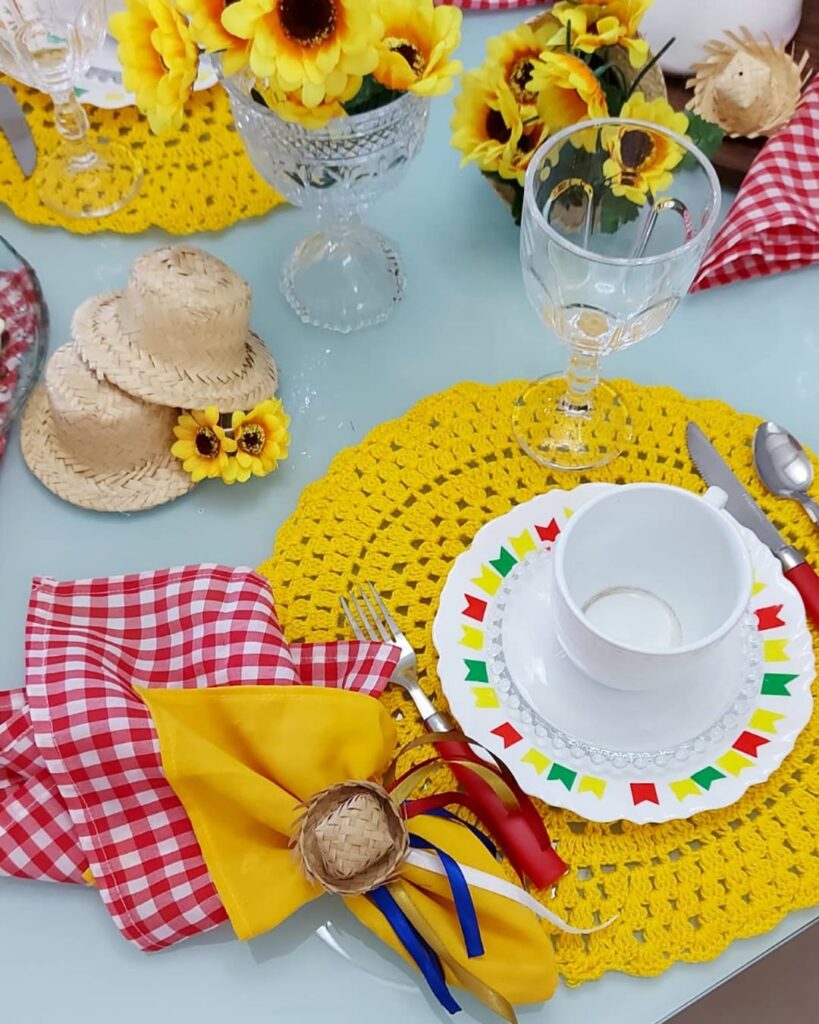Mesa posta de São João com sousplats de crochê amarelo, prato branco com bandeirinhas coloridas e guardanapo xadrez com chapéuzinho de palha