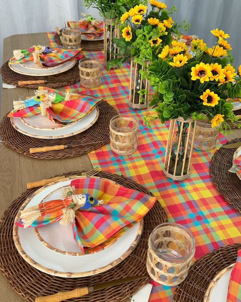 Mesa posta de São João com sousplats de vime, pratos brancos com detalhes na borda, caminho de mesa e guarnapos xadrezes com espantalhos