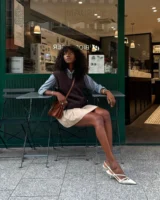 Mulher jovem de pele negra. Ela usa camisa, colete de tricô, saia e salto alto
