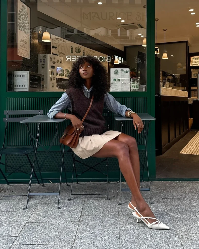 Mulher jovem de pele negra. Ela usa camisa, colete de tricô, saia e salto alto