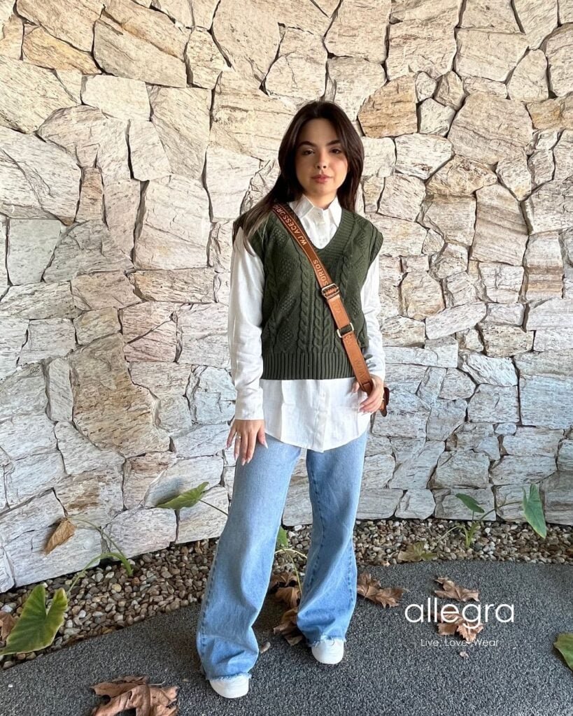Mulher jovem de pele clara. Ela usa camisa, colete de tricô, calça jeans e tênis branco