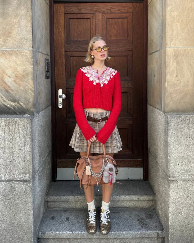 Mulher jovem, magra e de pele clara. Ela usa cardigan vermelho, minissaia xadrez plissada e tênis com meia