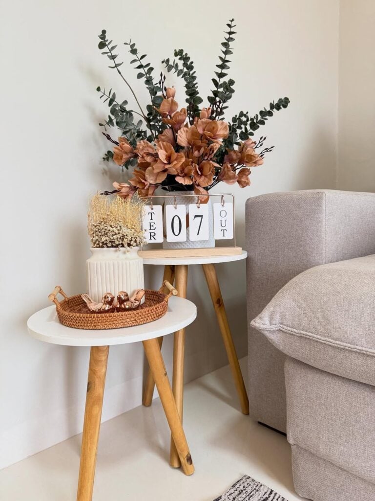 Mesinhas de sala com pés de madeira e tampos brancos com bandeja de palha e itens decorativos, e vaso com flores secas