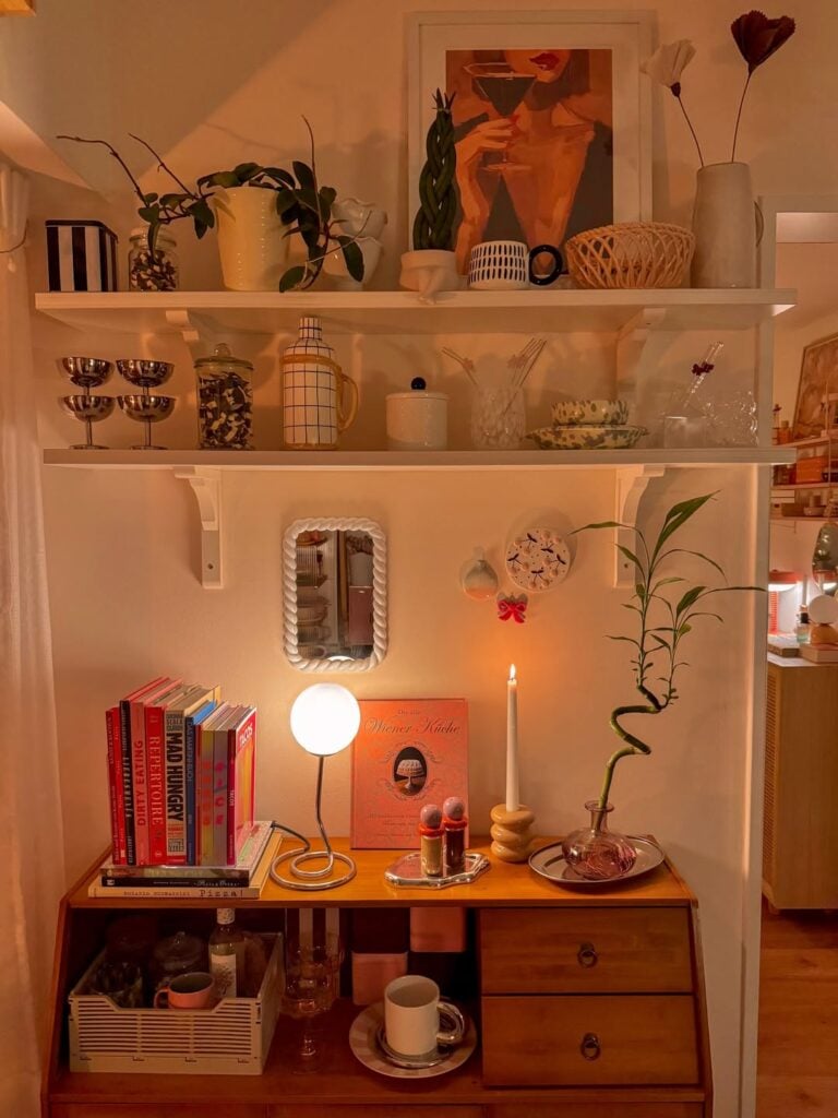Móvel de madeira com livros e luminária, e prateleiras brancas com objetos decorativos