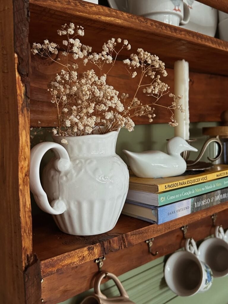 Prateleira de madeira com livros, jarra branca com florzinhas