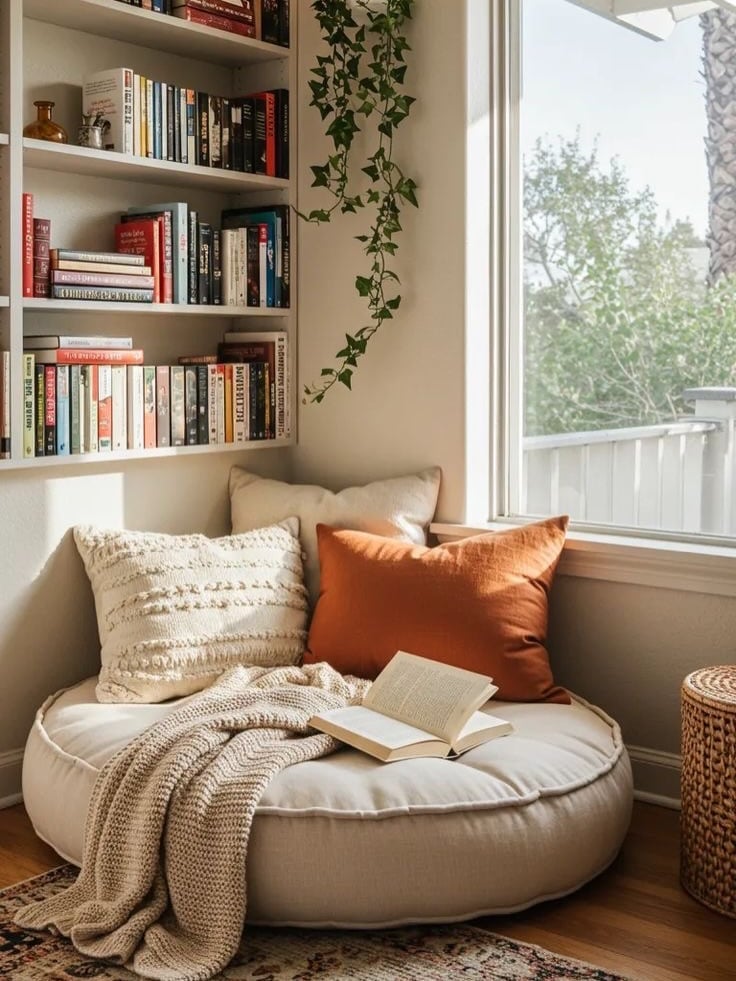 Espaço com pufe redondo claro, almofadas, manta e estante com livros