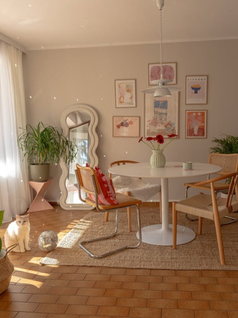 Sala de jantar com mesa redonda, cadeiras diferentes, quadros na parede e espelho com moldura orgânica