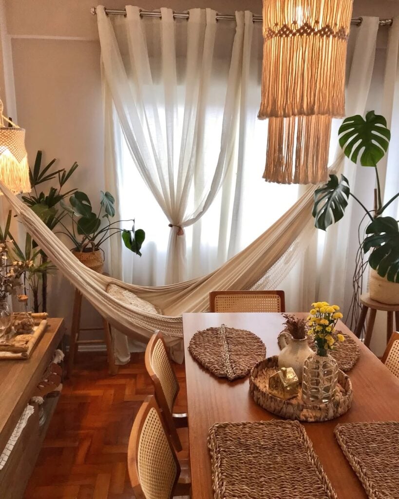 Sala de jantar com itens de palha na mesa de madeira, rede, plantas e cortina branca