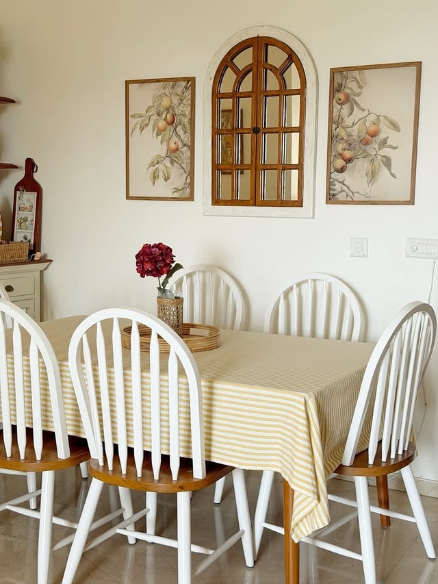 Mesa e cadeiras brancas, quadros com flores na parede e bandeja de palha com vaso e flores