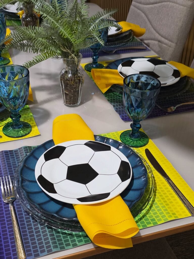 Mesa posta para Copa, com jogo americano colorido, pratos e taças azuis, e guardanapos amarelos
