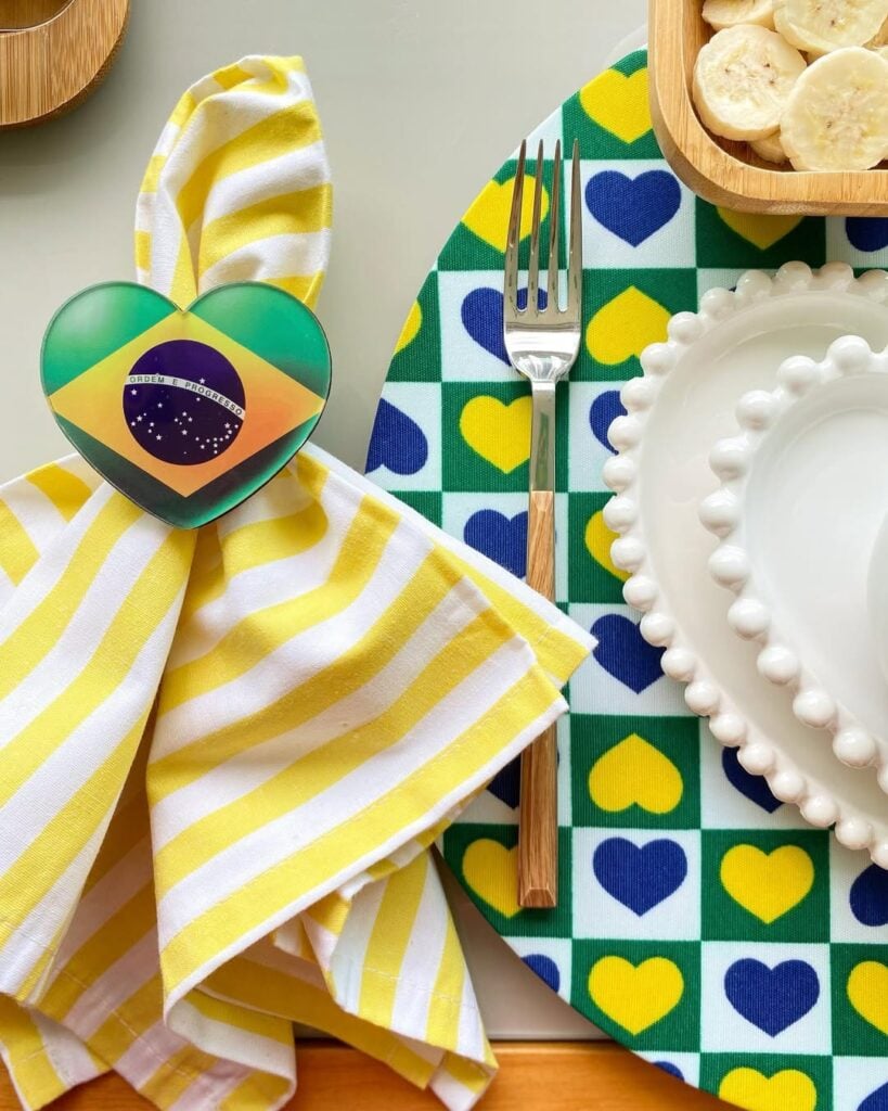 Mesa posta para a Copa com sousplat com corações azuis e amarelos e guardanapo listrado com coração e a bandeira do Brasil