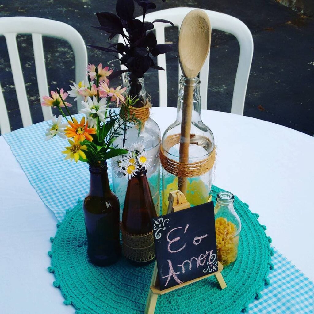 Mesa decorada para chá de cozinha com sousplat de crochê azul, garrafas de vidro com flores e colher de pau