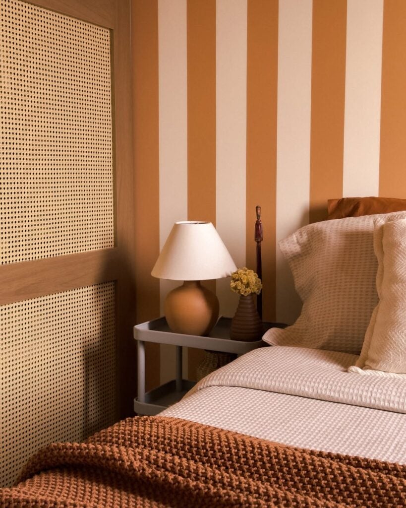 Quarto com parede listrada e carrinho ao lado da cama com abajur