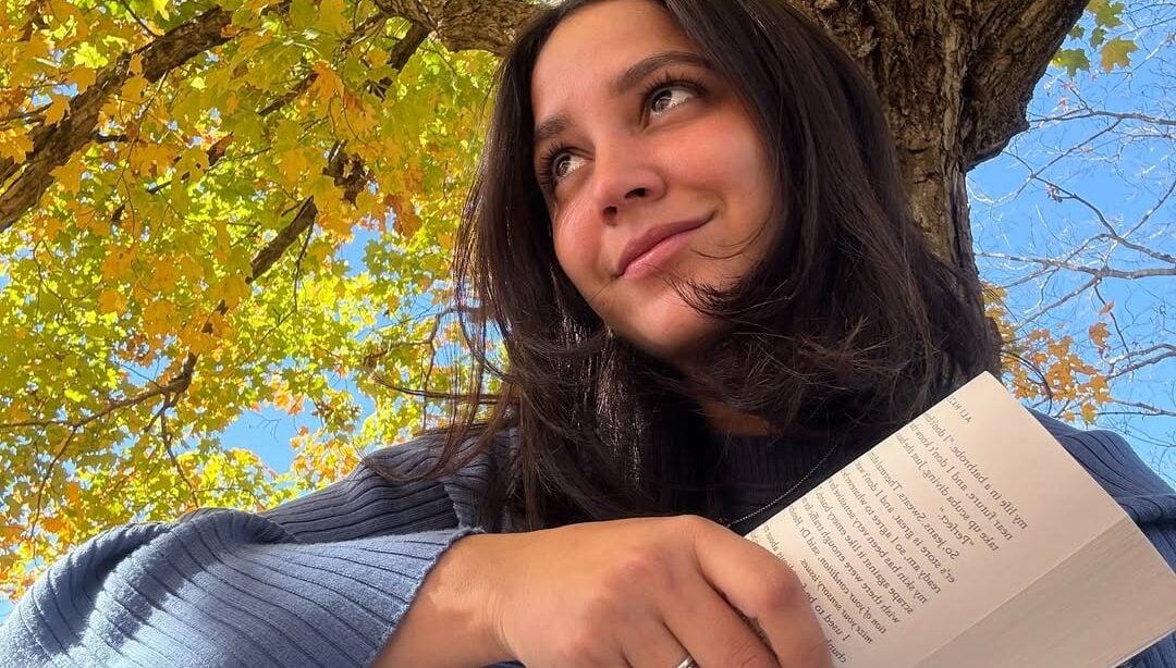 Mulher de pele clara, cabelo preto e look em tom de azul. Ela está sentada segurando um livro
