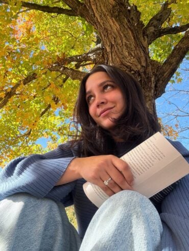Mulher de pele clara, cabelo preto e look em tom de azul. Ela está sentada segurando um livro
