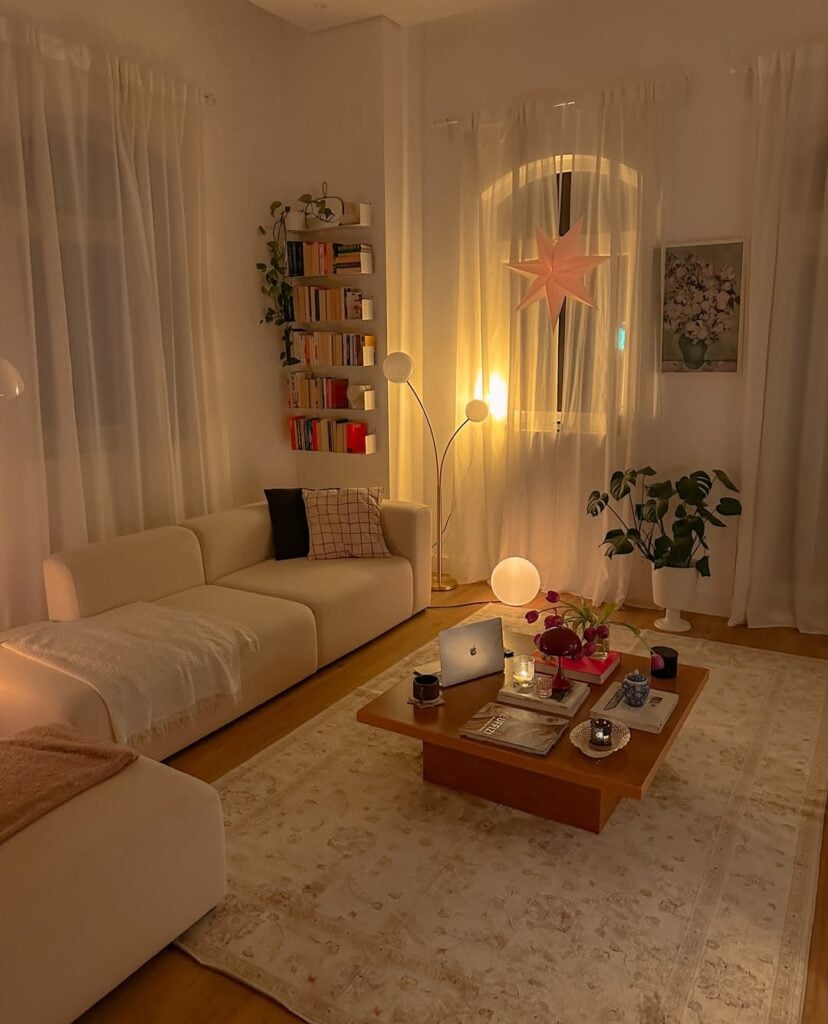 Sala de estar ampla com sofá branco, mesa de centro de madeira, cortinas e prateleiras com livros