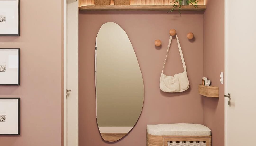 Hall de apartamento com a parede rosa, espelho orgãnico, banco e suporte para bolsas