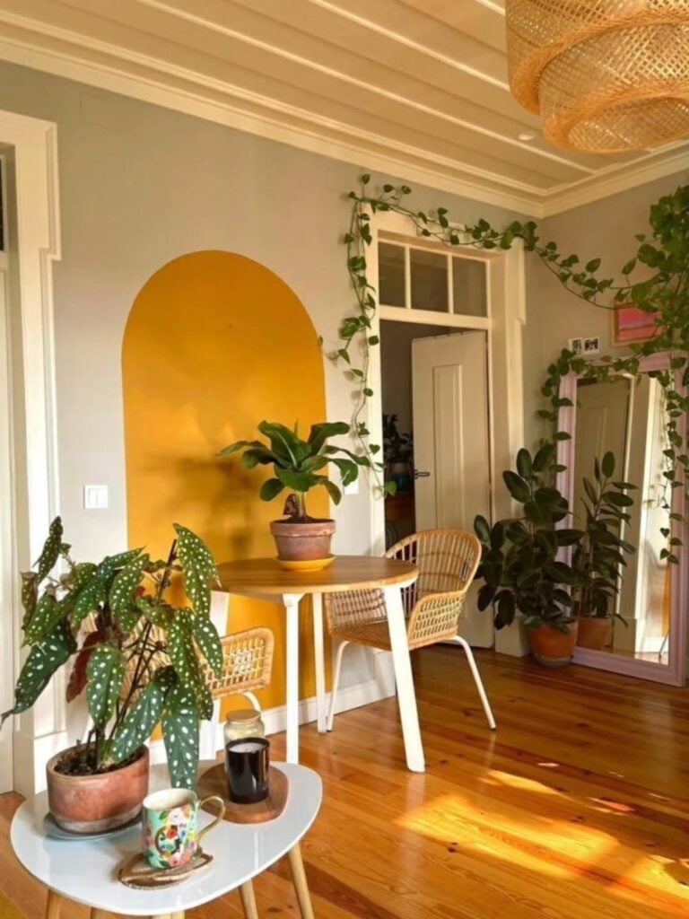 Ambiente com parede cinza e pintura orgânica amarela, mesa com cadeira de vime e plantas diversas