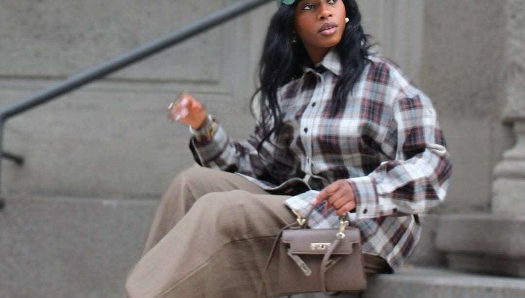 Mulher jovem com pele negra e cabelos escuros. Ela usa boné, camisa xadrez, calça e bota de cano curto e bico fino